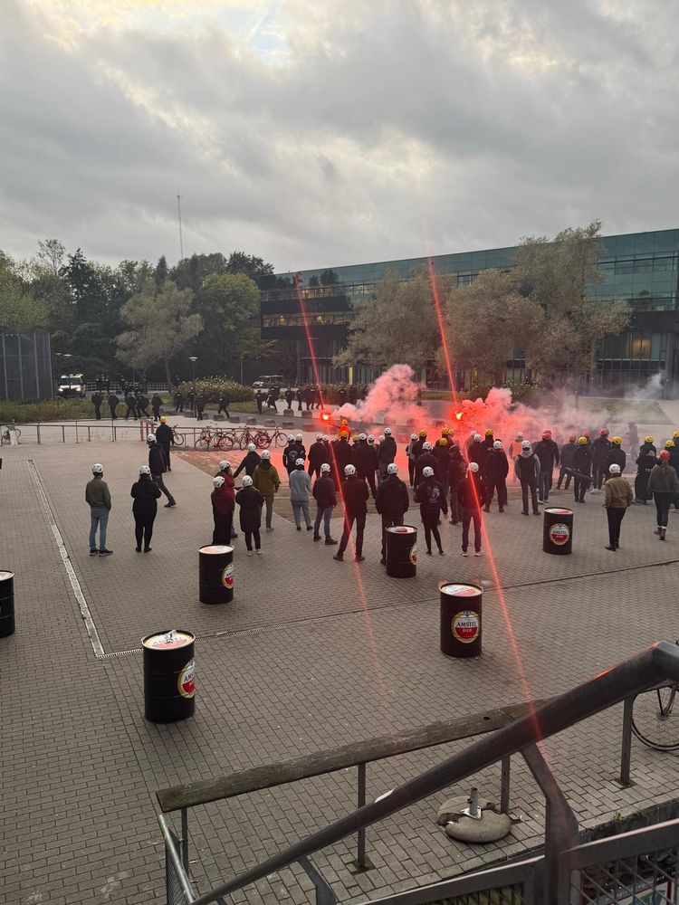 Tegenspelers met helm tijdens een groepsscenario met fakkels en rook, geobserveerd door beveiligers